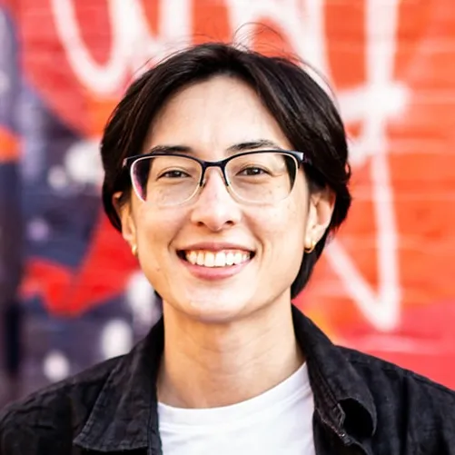 Headshot of Kai Wong. A nonbinary person smiling with short black hair, wearing glasses, a black button up and white tee shirt against a colorful background.